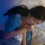 A woman lying in bed at night, holding her chest and coughing, struggling to stop coughing and find relief for a restful sleep.