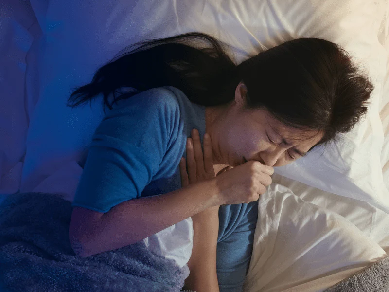 A woman lying in bed at night, holding her chest and coughing, struggling to stop coughing and find relief for a restful sleep.