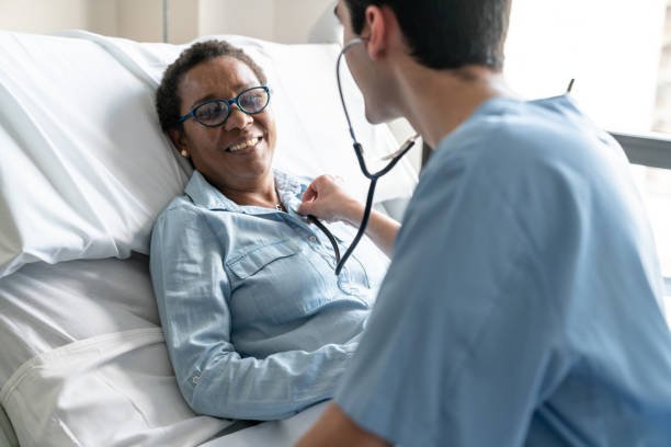 Female black friendly patient lying down on hospital bed while nurse is checking her heart beat with stethoscope